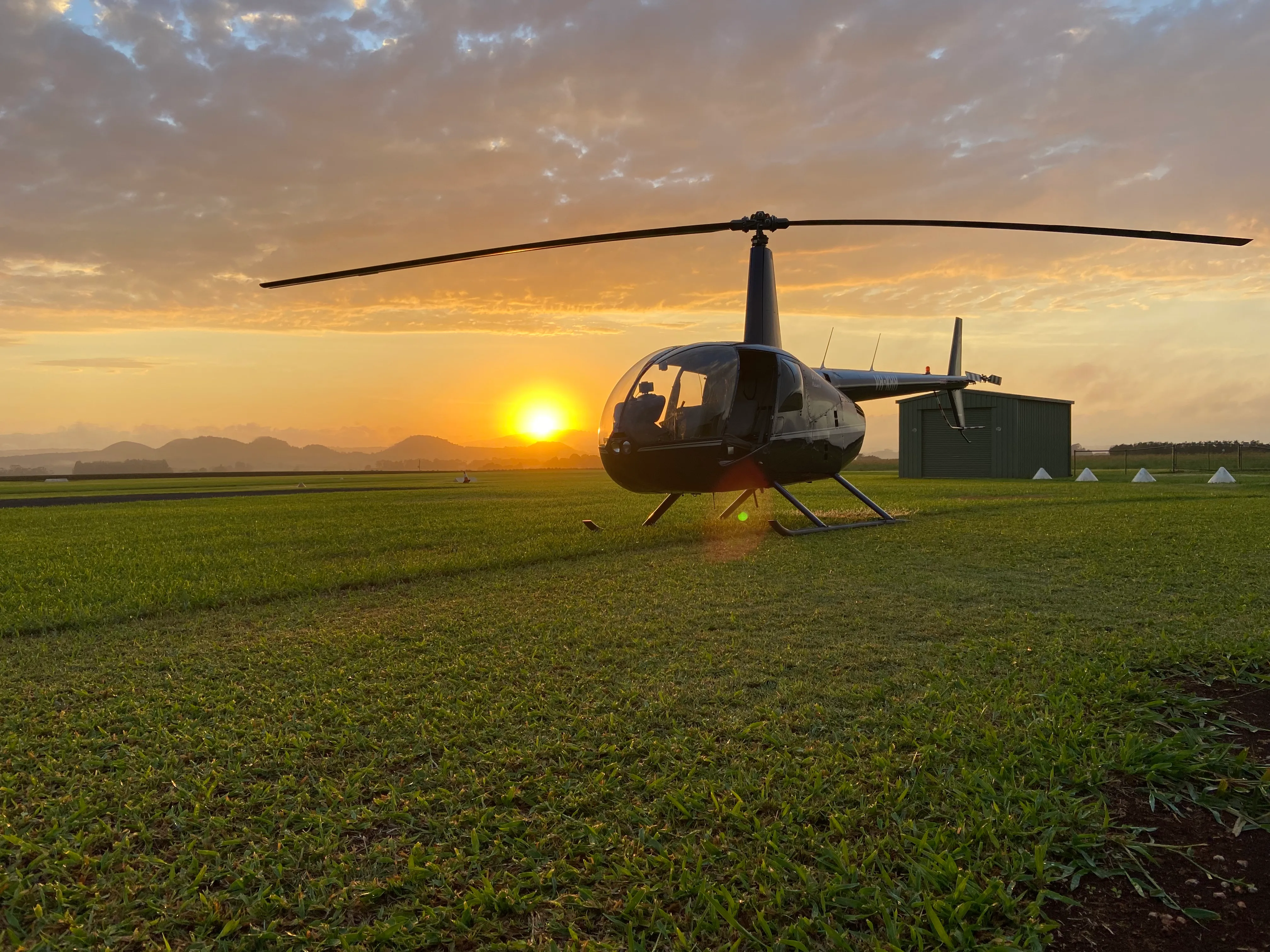 A helicopter on the ground at sunset with a hangar and hills in the background.