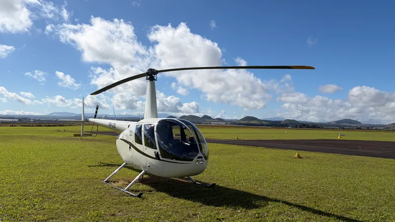 An R44 helicopter parked on the grass at Atherton Aerodrome under a blue sky, with the Tablelands hills in the distance.